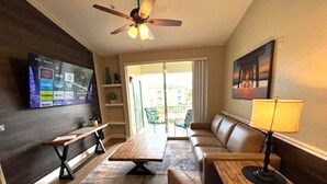 Une chambre d’hôtel comprenant un canapé, une table en bois, un banc, un téléviseur fixé au mur et un ventilateur de plafond.