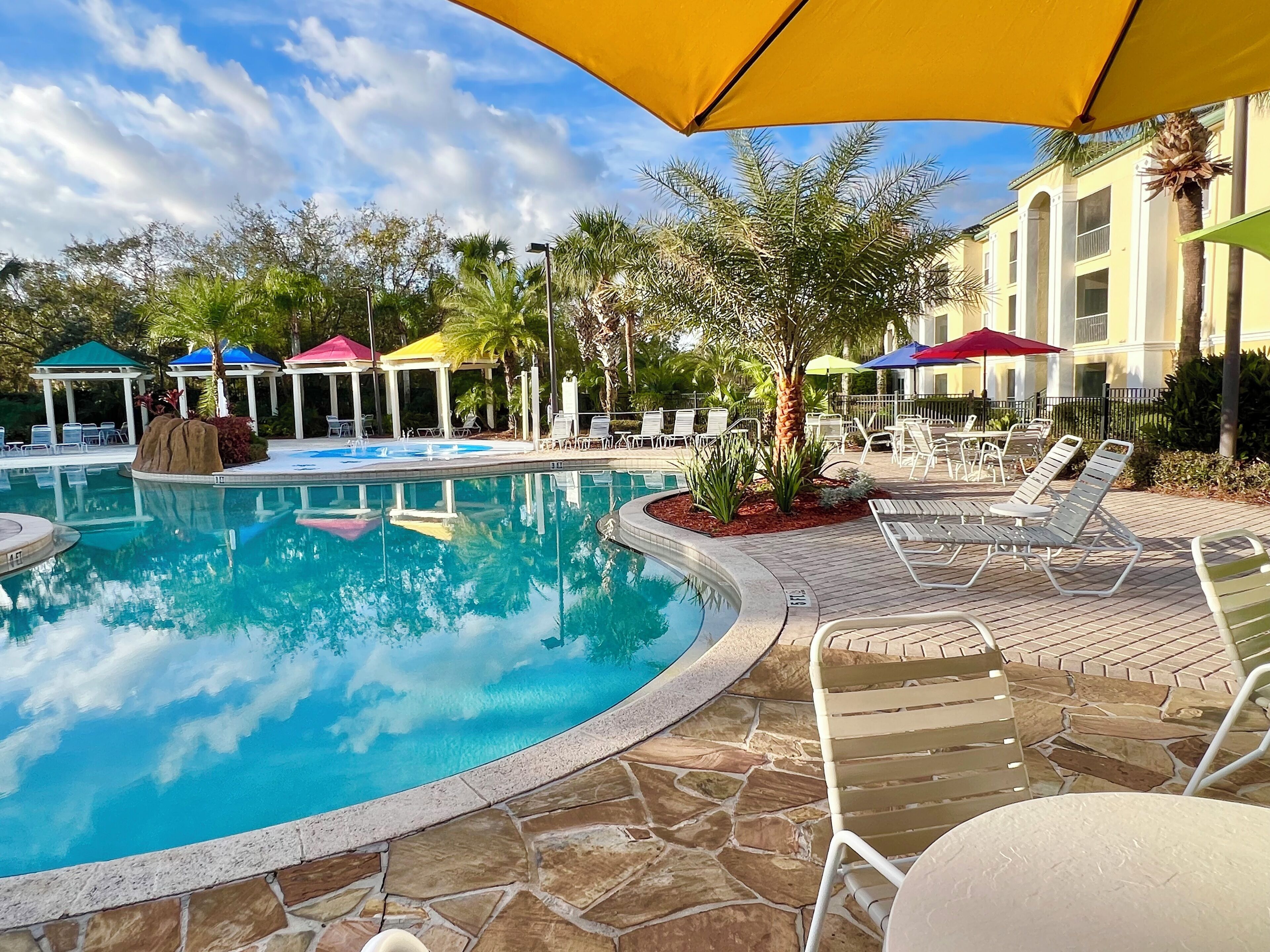 A pool area with lounge chairs and umbrellas, surrounded by a stone patio and greenery.