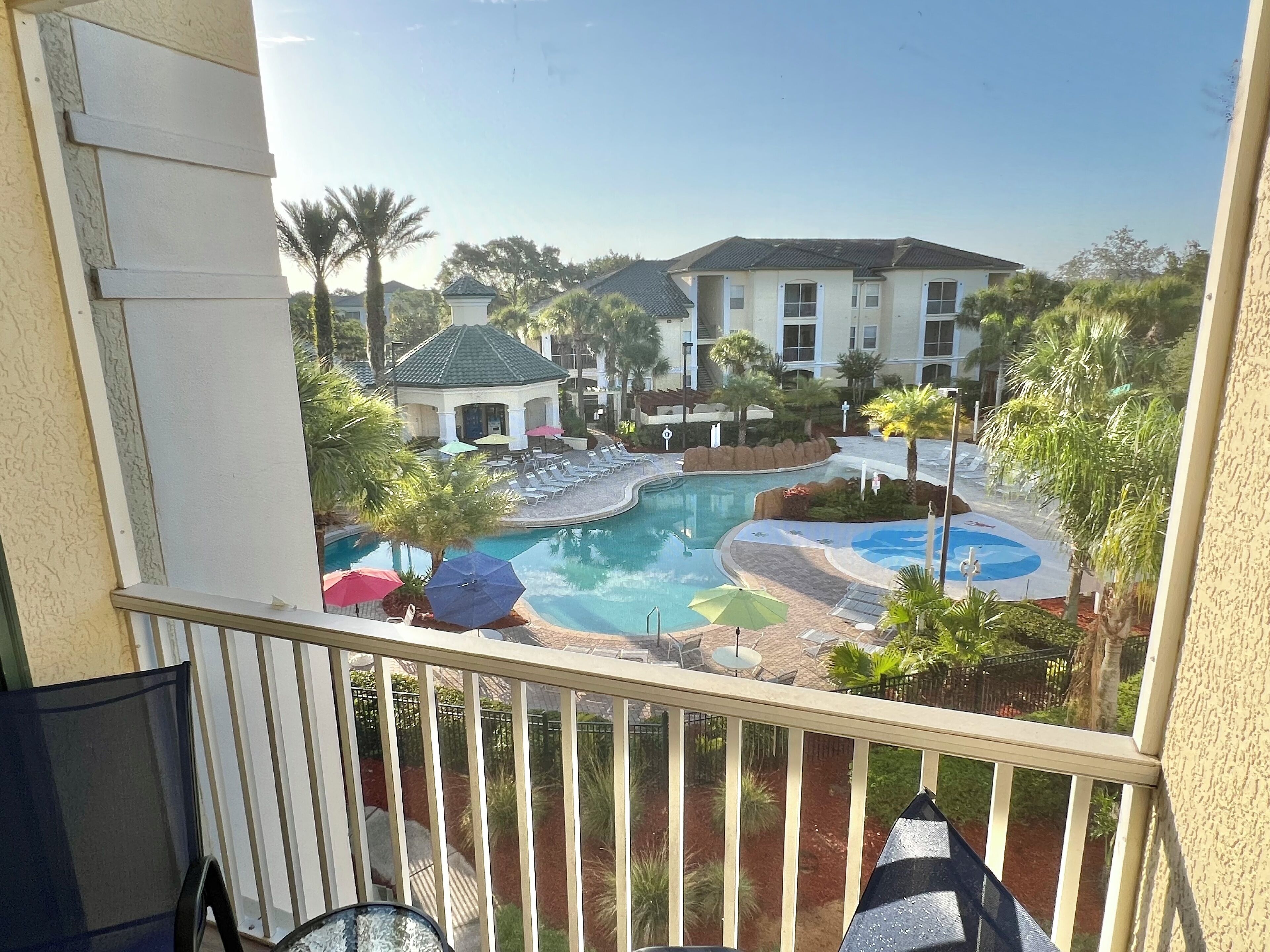 A pool area with lounge chairs and umbrellas, surrounded by palm trees and a building with a tiled roof.