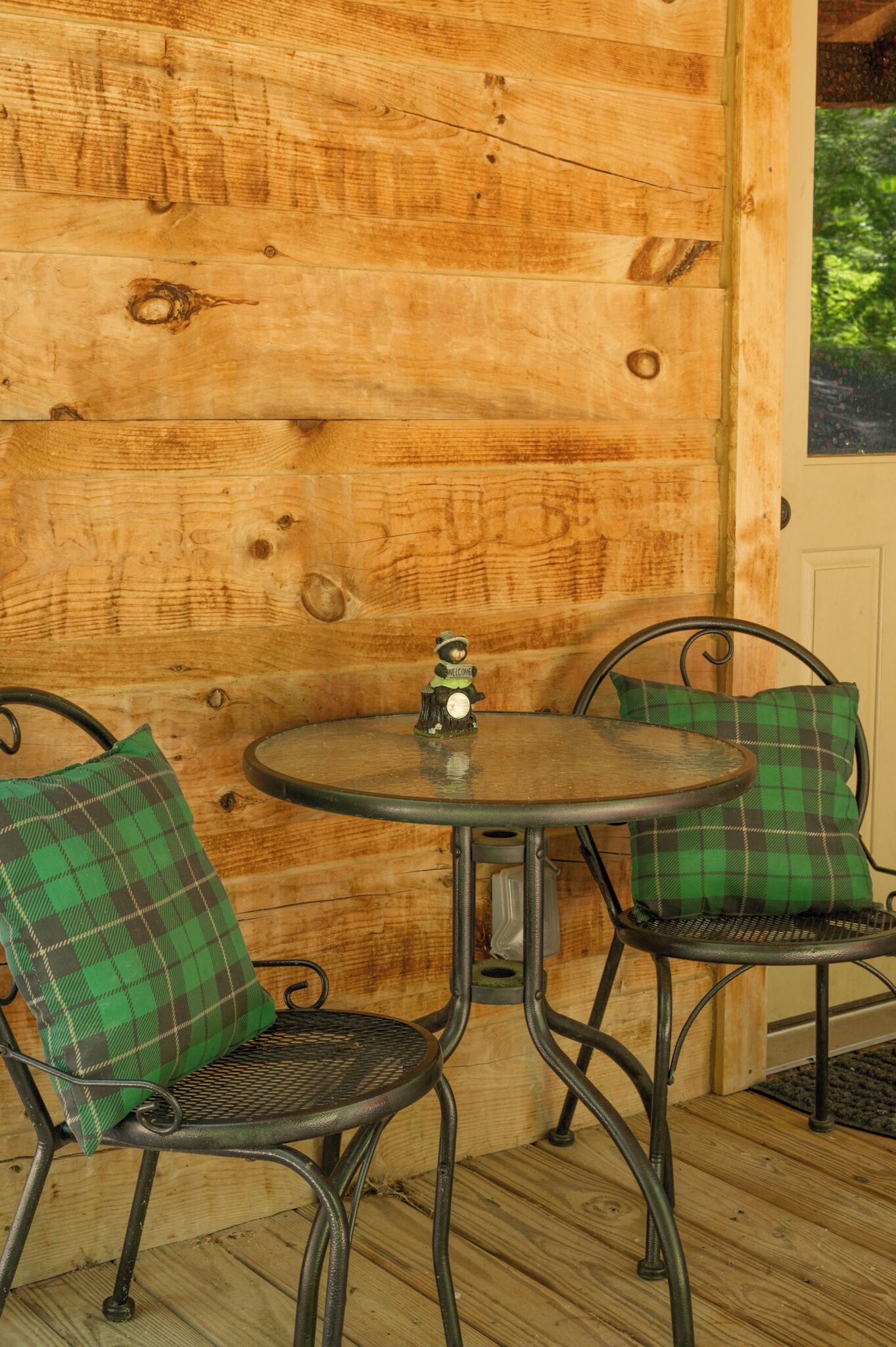 A wooden wall with a round table and two chairs, each with a green plaid cushion.