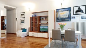 Una sala de estar moderna con suelo de madera, un sofá blanco, una mesa de comedor con sillas, un televisor y una estantería.