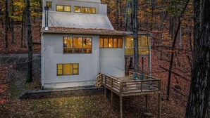 Una cabaña moderna con terraza de madera y varios ventanales, rodeada de un bosque.