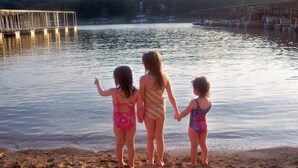 Drie kinderen staan hand in hand op een zandstrand, kijkend naar een kalm meer met een pier op de achtergrond.