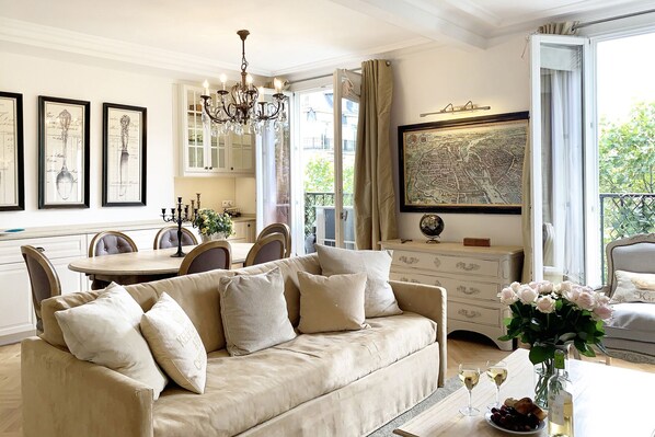 A spacious living room with a beige sofa, a dining area with a chandelier, and a large window with a cityscape view.