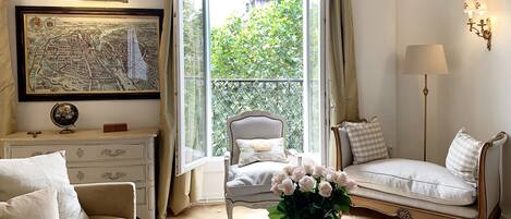 A well-lit room with a large window, a framed map, a globe, a sofa, a chair, and a table with a vase of flowers and a bottle.