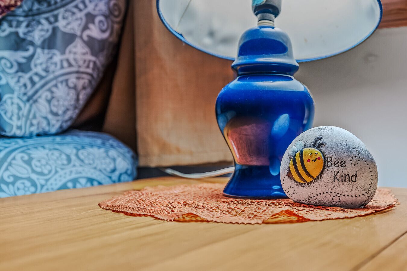 A blue lamp, a white rock with a bee drawing and text, and a patterned cushion on a wooden table.