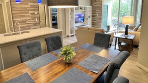A modern kitchen with a central island, dining area with a wooden table and chairs, and a living room with a sofa and a TV.