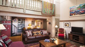 A living room with a brown leather sofa, a wooden coffee table, a red armchair, a television, and a colorful hanging tapestry. - Spacious, Sunlit Adobe Casita #6 / B&B in the Heart of Historic Taos! (Taos)