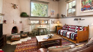 A cozy living room with a fireplace, wooden furniture, and a large window with a view of trees. - Spacious, Sunlit Adobe Casita #6 / B&B in the Heart of Historic Taos! (Taos)