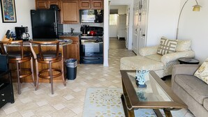 A modern kitchen with wooden cabinets, a black refrigerator, a microwave, and a stove. There is a dining area with a bar stool and chairs. A living room with a sofa and a coffee table is visible.