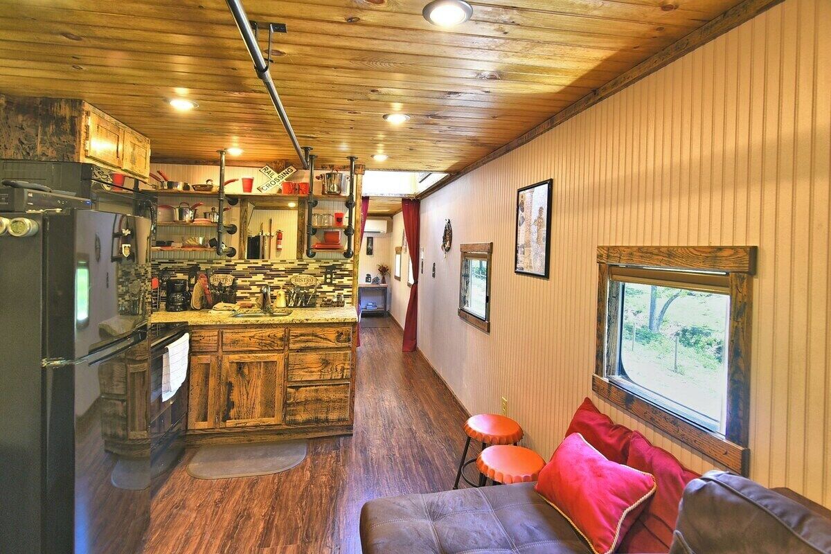 Una cocina compacta con armarios de madera y una isla central, un comedor con un banco y cojines rojos, y una ventana con vistas a zonas verdes.