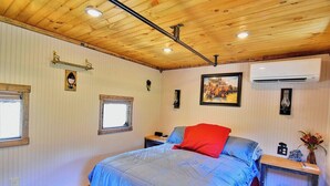 A bedroom with a wooden ceiling, a bed with a blue blanket, a red pillow, a nightstand with a radio, and a wall-mounted air conditioner.