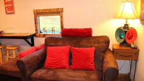 A cozy room with a brown sofa, red cushions, a wooden table, a lamp, and a window with a view of deer.