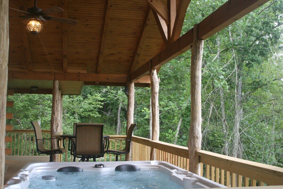 A hot tub on a wooden deck with chairs, surrounded by trees.