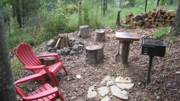 A forested area with a red Adirondack chair, a fire pit, a stack of firewood, and a grill.