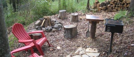 A forested area with a red Adirondack chair, a fire pit, a stack of firewood, and a grill.