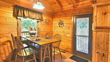 A wooden interior with a dining table, chairs, a window with a view, and a door leading outside.