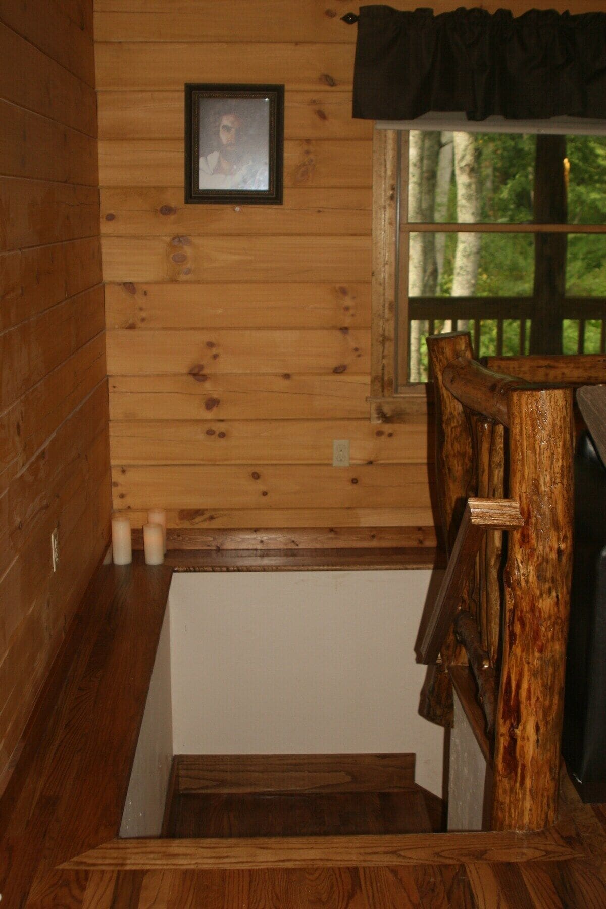 A wooden staircase with a wooden railing, a window with a view of trees, and a framed picture on the wall.
