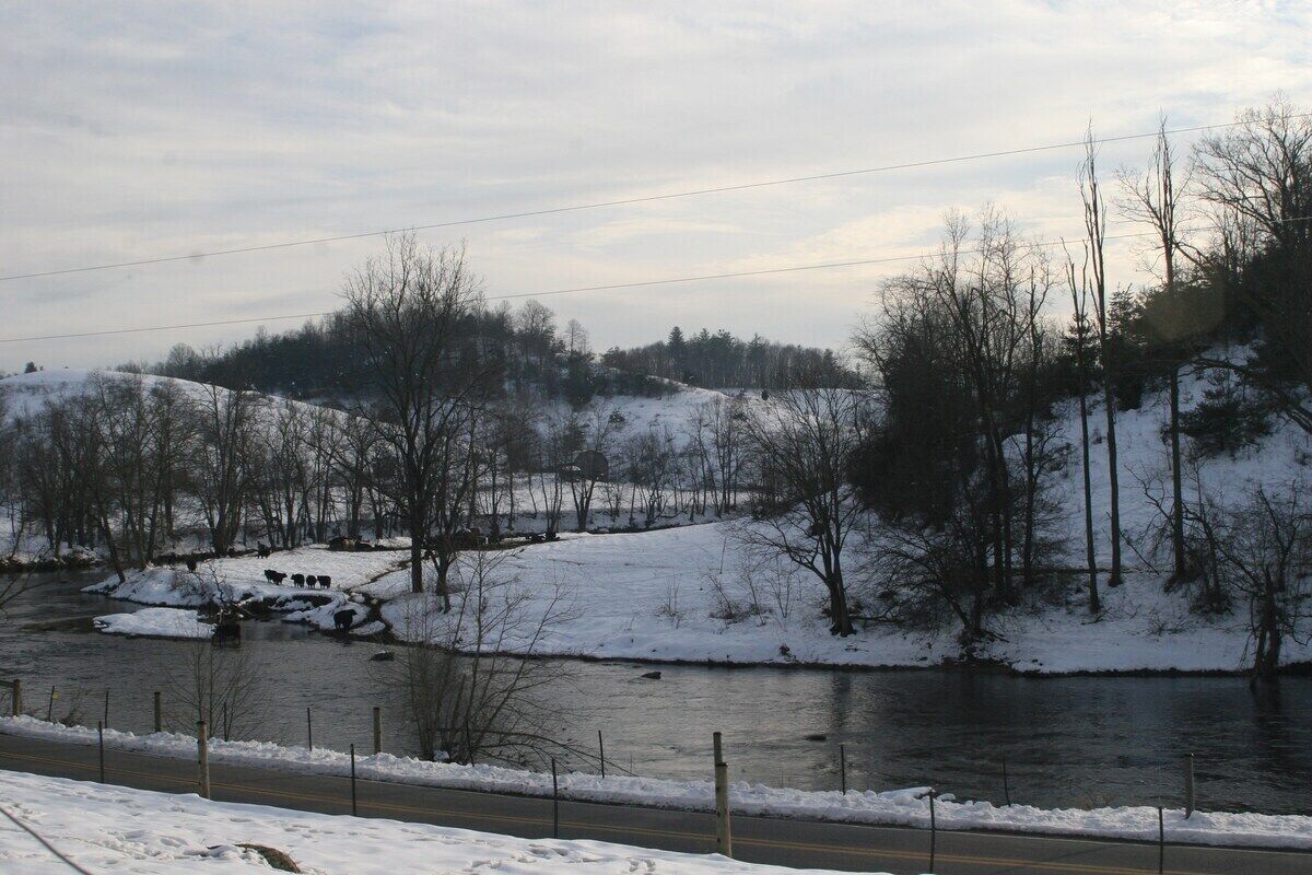 A snow-covered landscape with a river, trees, and a road.