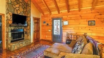 A log cabin interior with a stone fireplace, a television mounted above it, a wooden ceiling, and a patterned rug.