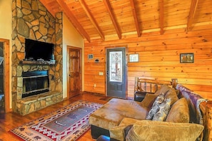 Ein rustikales Blockhaus-Interieur mit Kamin aus Naturstein, einem darüber angebrachten Fernseher, einer Holzbalkendecke und einem gemusterten Teppich.