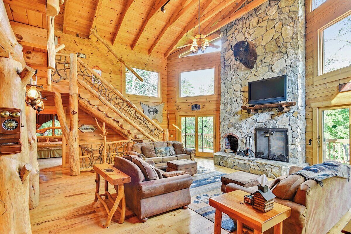 A log cabin interior with a stone fireplace, wooden staircase, and a television mounted on the wall.