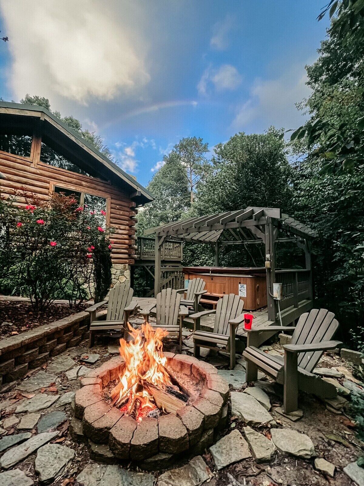 A fire pit with seating, surrounded by a wooden pergola, set in a garden with trees and a wooden cabin in the background.