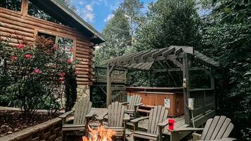 A fire pit with seating, surrounded by a wooden pergola, set in a garden with trees and a wooden cabin in the background.