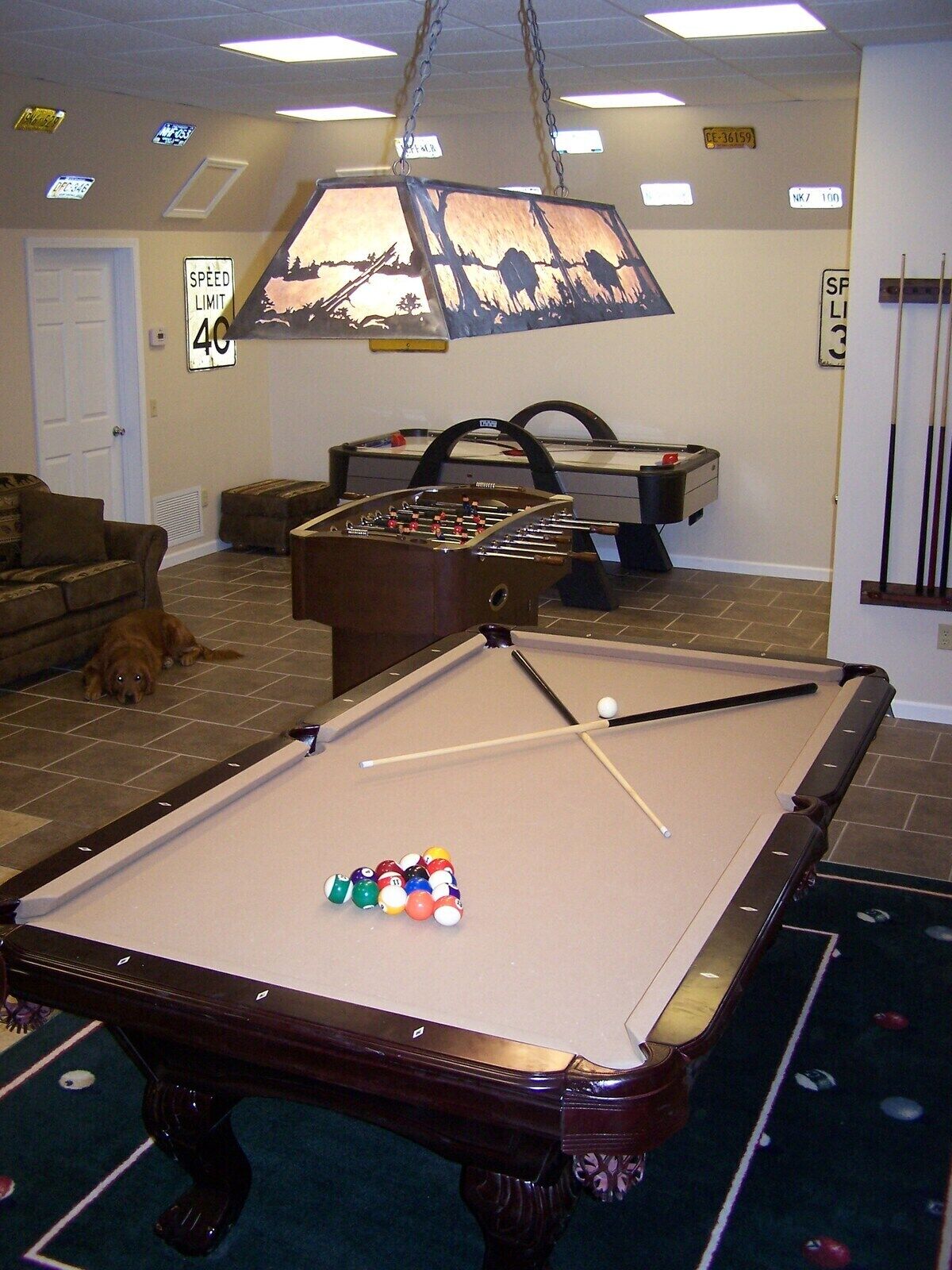 A pool table with balls, a foosball table, and a dog in a room with a ceiling light fixture.