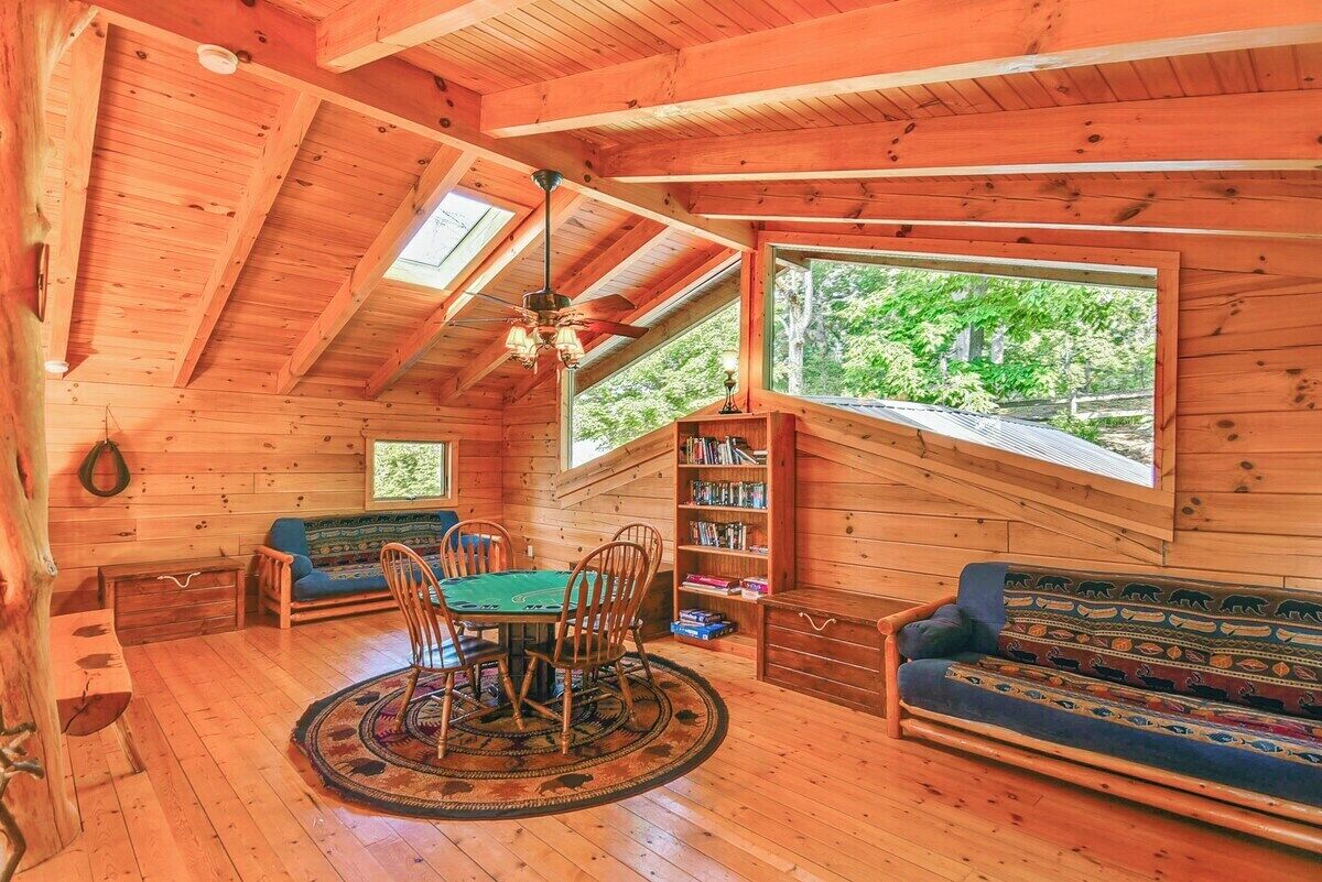 A wooden cabin interior with a dining table, chairs, a sofa, and a bookshelf.