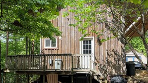 A rustic wooden cabin with a deck, two Adirondack chairs, and a stone wall.