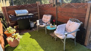 Ein Garten mit Grill, zwei Stühlen, einem kleinen Tisch mit einem Getränk und Topfpflanzen.