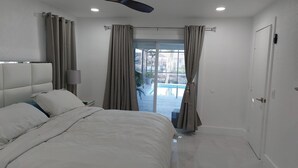 Ein Schlafzimmer mit Bett, Nachttisch, Lampe und Blick auf einen Pool durch Vorhänge.