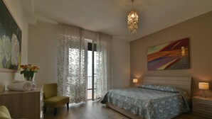 Un dormitorio con una cama, una silla, un escritorio con un florero y un cuadro en la pared.