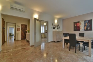 A spacious living area with a dining table and chairs, a kitchen in the background, and a hallway leading to other rooms.