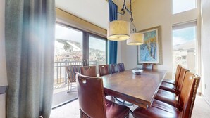 Un comedor con una amplia mesa de madera y sillas de cuero, ventanas grandes que ofrecen vistas a un paisaje nevado y una lámpara colgante.