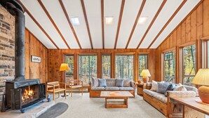 A cozy cabin interior with a fireplace, wooden furniture, and large windows offering a forest view. - Haven Home - deck, grill, wifi, pool access, hiking, wood stove, games (Berkeley Springs)