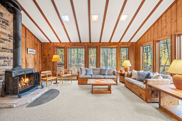 A cozy cabin interior with a fireplace, wooden furniture, and large windows offering a forest view.