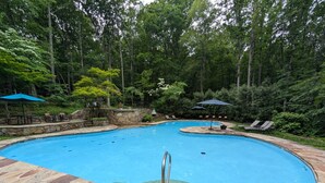 A swimming pool with a stone deck, surrounded by trees and shaded areas with umbrellas. - Haven Home - deck, grill, wifi, pool access, hiking, wood stove, games (Berkeley Springs)