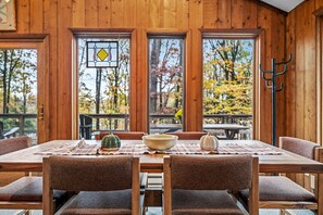 Une salle à manger en bois, avec une table dressée pour quatre personnes, de grandes fenêtres et une vue sur les arbres aux couleurs automnales.