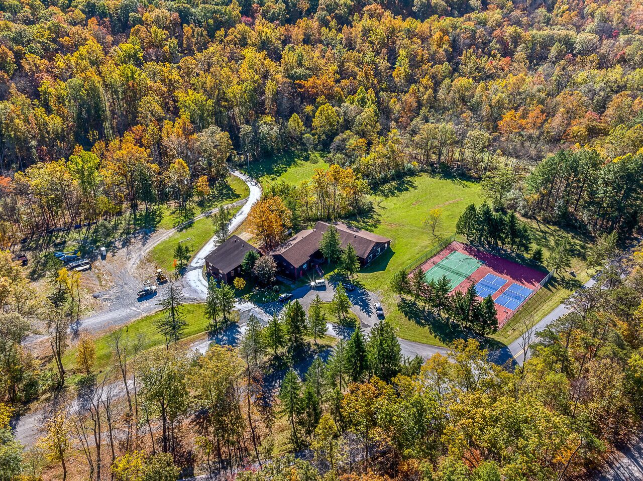 An aerial view of a property with a large building, a tennis court, and surrounding wooded area.