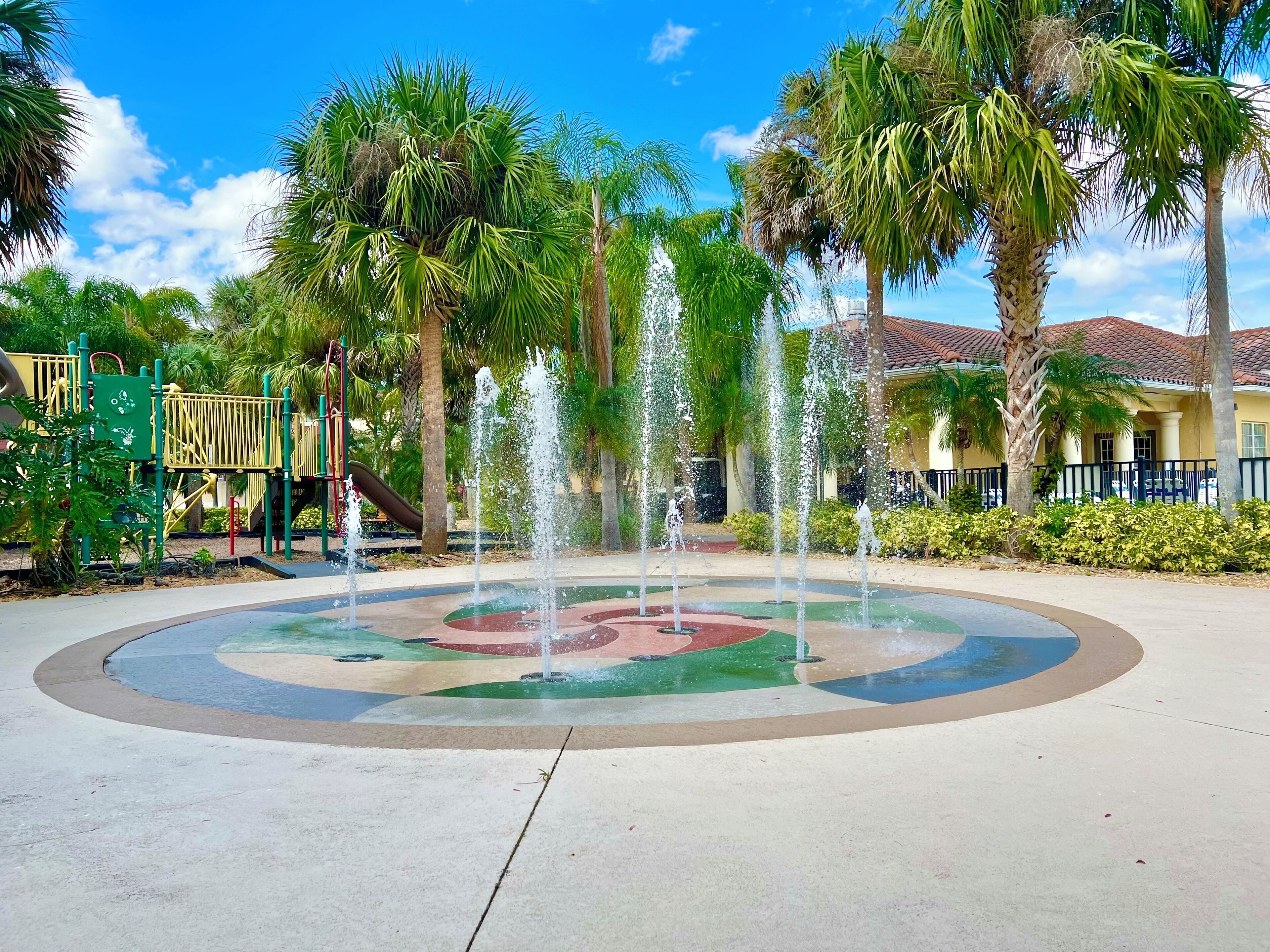 Une fontaine avec plusieurs jets d’eau disposés en cercle, entourée de palmiers et d’une aire de jeux avec un toboggan.