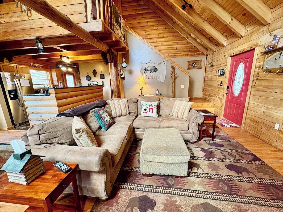 A cozy cabin interior with a sectional sofa, a small ottoman, a wooden coffee table, and a red door.