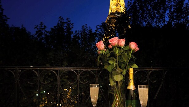 A table with a bottle of champagne, a glass, and a vase with roses against a night sky.