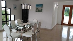 Un comedor moderno con una mesa de vidrio y sillas blancas, un mueble negro y un cuadro con temática playera en la pared.