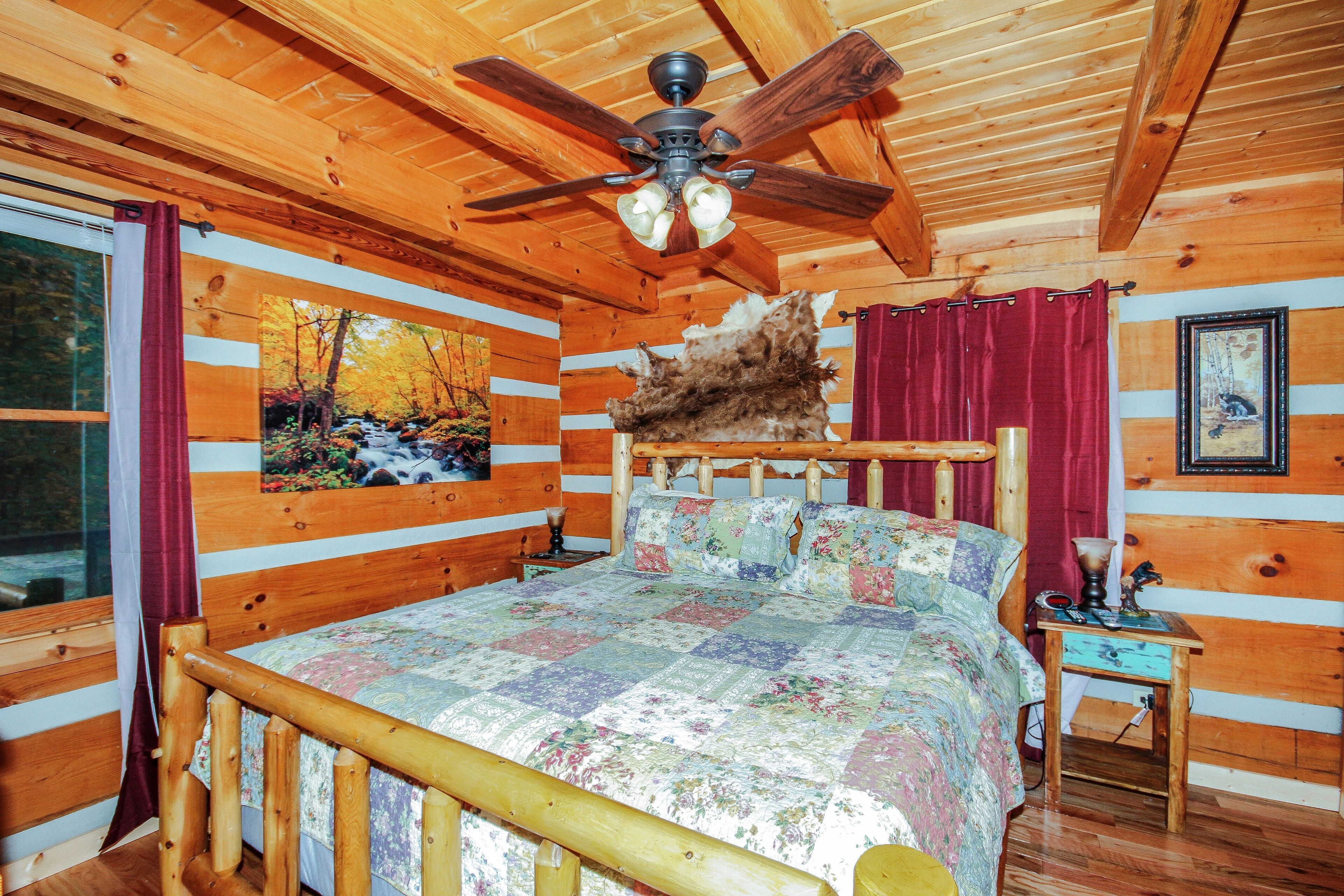 A wooden cabin bedroom with a bed, a ceiling fan, a painting of a forest, a fur rug, and a wooden nightstand.