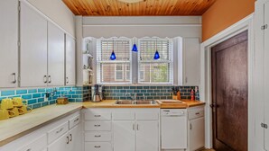 Une cuisine avec des meubles blancs, un plan de travail en bois, un évier et deux fenêtres ornées de suspensions lumineuses bleues.