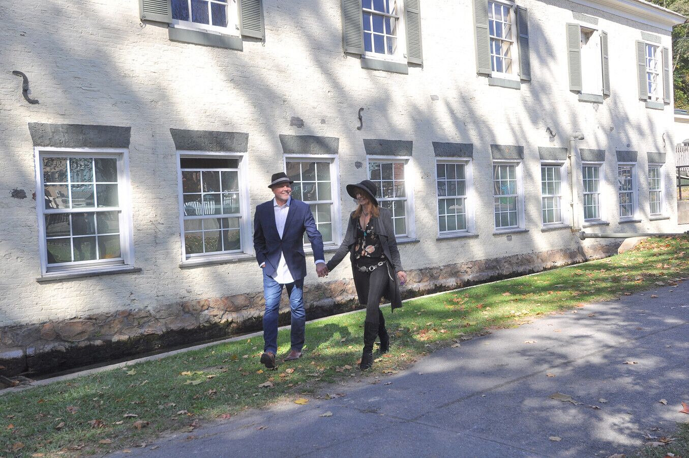 A man and woman holding hands walking on a sidewalk next to a building with multiple windows and a stone foundation.