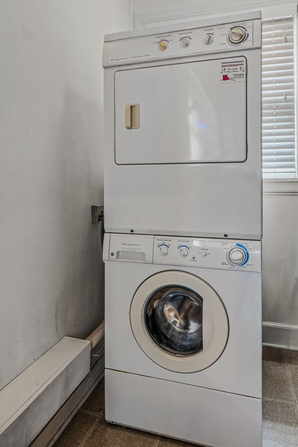 Lavadora y secadora apiladas en un rincón de una habitación con piso de baldosas y una ventana con persianas.