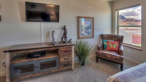 A room with a large TV mounted on the wall, a framed painting, a wooden desk with drawers, a leather armchair with colorful cushions, and a view of a mountainous landscape through a window. - DW6  Private Hot Tub, En-Suite Bathroom for Each Bedroom, Near Arches Park! (Moab)